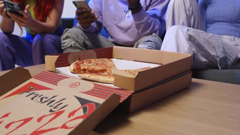 close up of gen z friends sitting on sofa eating home delivery takeaway pizza and using mobile phones