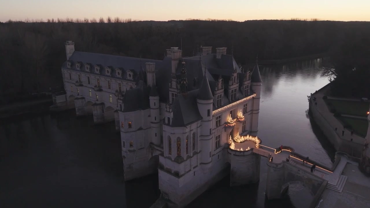 Une vue aérienne au crépuscule montre le Château de Chenonceau à côté de son jardin à la française, s'illuminant doucement dans la lumière du soir