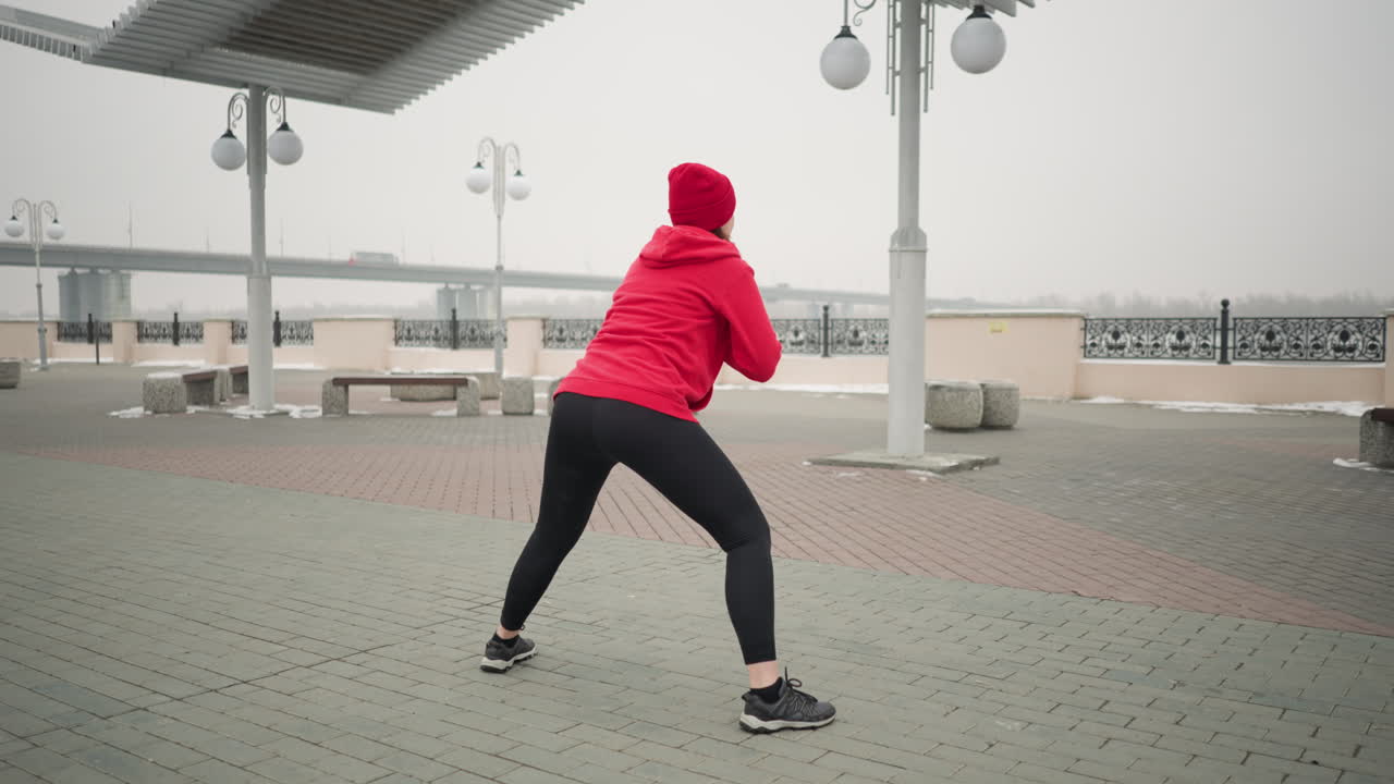 Back view of woman stretching outdoors in winter, performing side lunges on paved ground under lamp posts and pergola near bench and railing, winter workout scene with bridge and foggy atmosphere