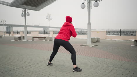 back view of woman stretching outdoors in winter, performing side lunges on paved ground under lamp posts and pergola near bench and railing, winter workout scene with bridge and foggy atmosphere