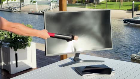 a person uses a hammer to destroy a computer monitor on a patio table beside a river or canal, under bright natural daylight with steady camera framing