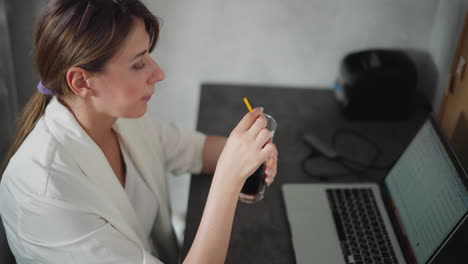focused data analyst in white top working in home office sipping coffee from glass through yellow straw while checking spreadsheet on laptop sitting at gray desk with organized workspace