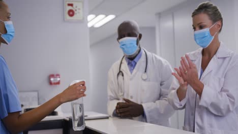 Diverse-hospital-worker-disinfecting-hands-of-doctors-all-wearing-face-masks