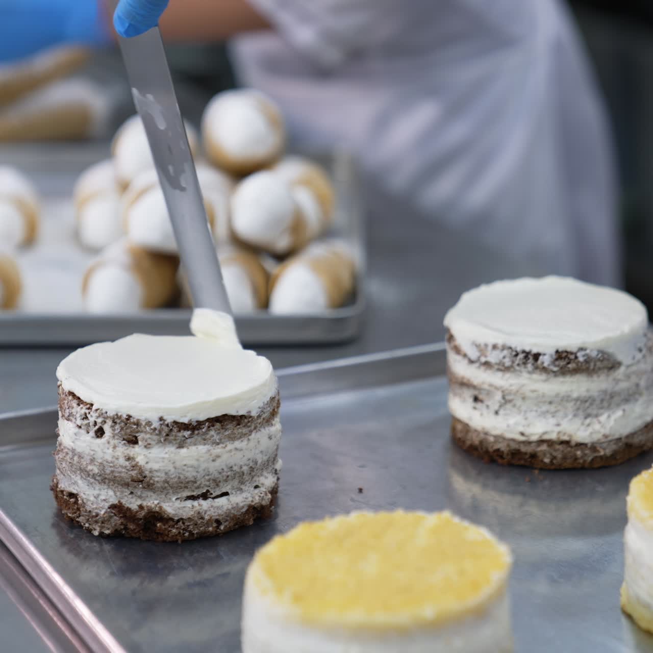 Worker in blue gloves covers little cakes with thick layer of cream. Confectioner uses spatula to decorate desserts. Blurred backdrop