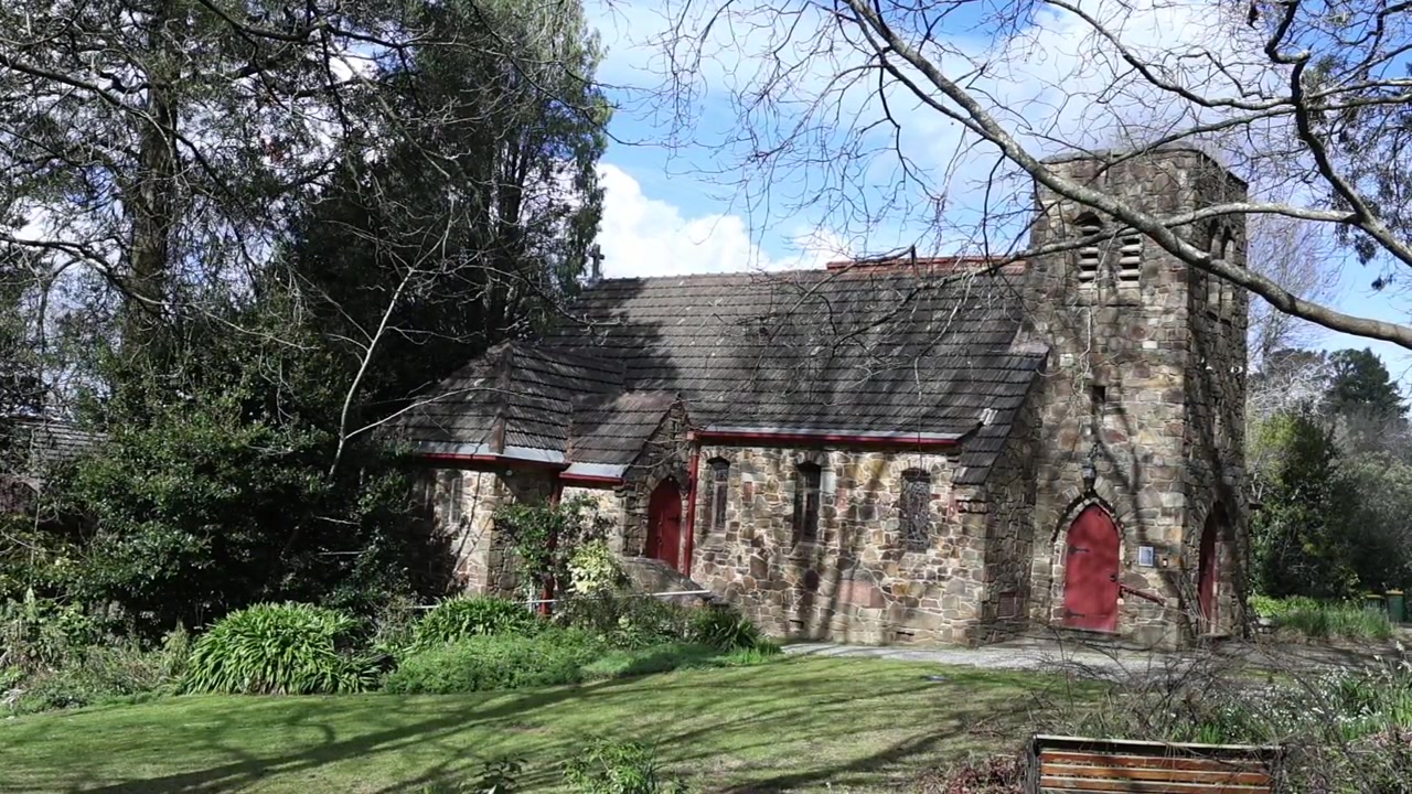 Vista grandangolare di una vecchia chiesa in pietra a Olinda, Melbourne, immersa tra giardini verdi e alberi alti sotto la luce diurna intensa. Architettura classica, atmosfera tranquilla, nessuna persona visibile.