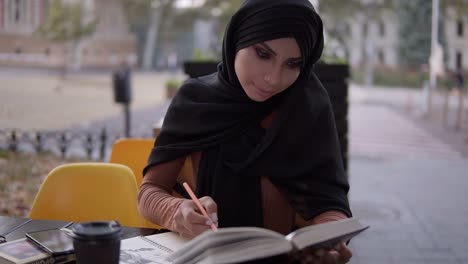 an attractive muslim girl writes in a notebook sitting in a cafe with a book. studying in outdoors cafe, having break. college, self education. front view. slow motion