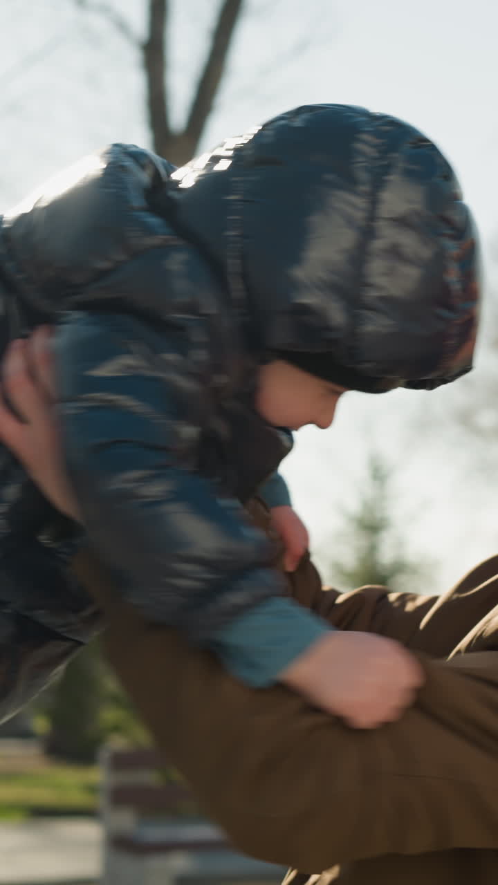 un primer plano de un padre y un hijo disfrutando del tiempo de juego al aire libre mientras el padre gira su hijo suavemente en un parque y lo coloca en sus hombros, con árboles y bancos en el fondo