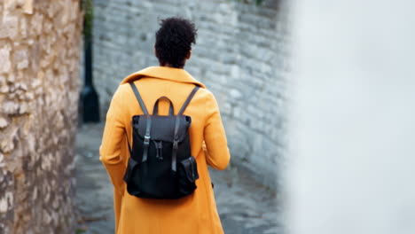 Back-view-of-young-woman-wearing-a-yellow-coat-walking-away-from-camera-down-an-alleyway-between-stone-walls,-selective-focus