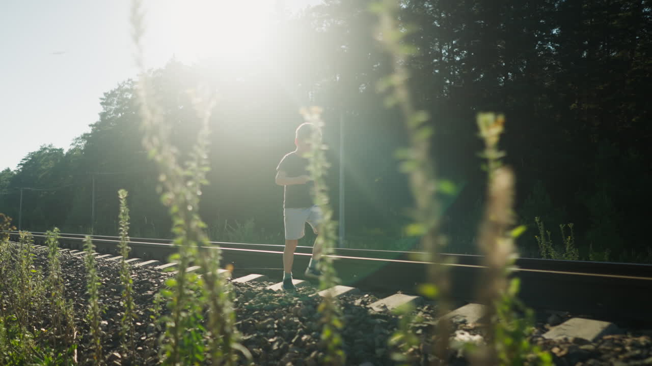 Little boy walks along rail track as sunlight creates dreamy glow around him, soft focus foreground plants frame scene, with deep forest shadows and power lines