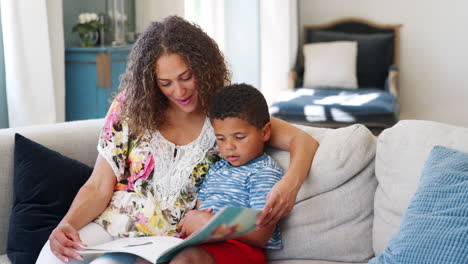 Toma-En-Cámara-Lenta-De-Una-Madre-Sentada-En-Un-Sofá-En-Casa-Leyendo-Un-Libro-Con-Su-Hijo