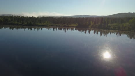 aerial shot panning left over a calm watered lake with sun reflected facing a beautiful forest in northern ontario canada