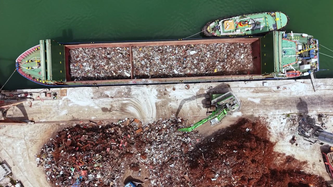 Nave che scarica materiali vicino a un deposito di rottami in un porto durante il giorno, con macchinari e cumuli di metallo e detriti visibili nell'area.