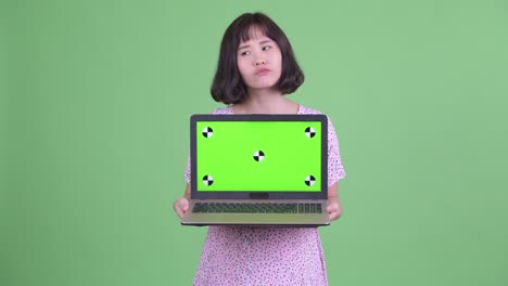 studio shot of beautiful pregnant asian woman showing laptop computer with copy space while thinking against green studio background