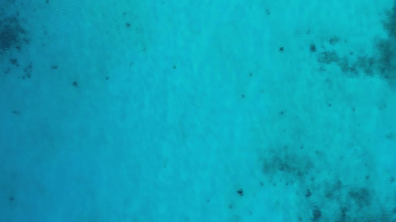 La arena blanca brilla bajo las aguas poco profundas del océano, mientras el dron asciende al cielo.