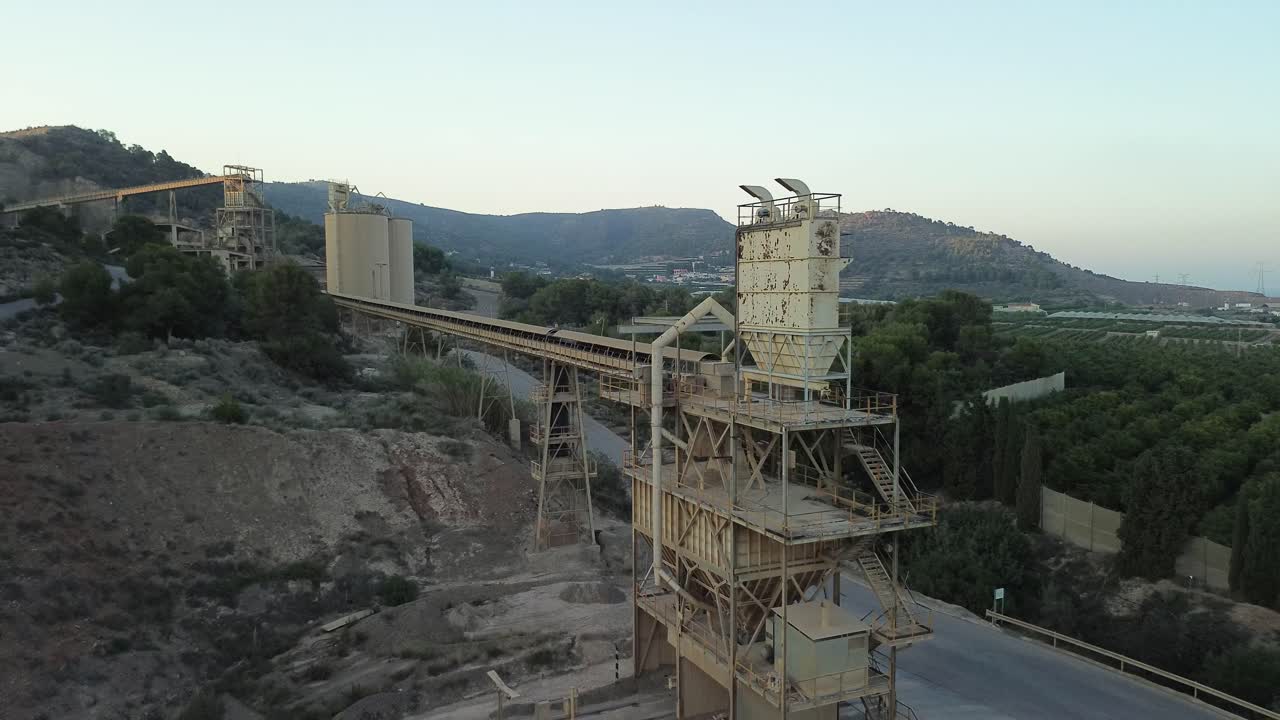 Industrial quarry with conveyor belts and stone crusher amidst hills, showcasing heavy machinery and mining operations. Drone shot moving away