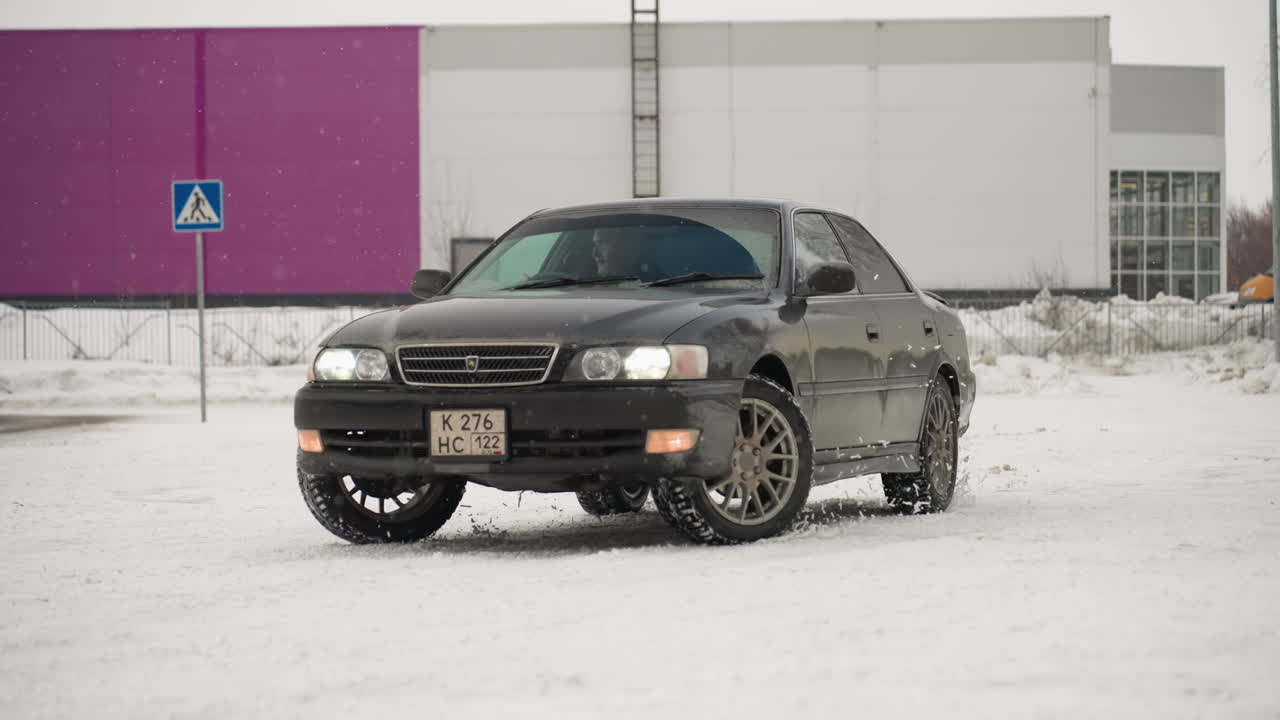 car making controlled turn on snowy road in winter near pedestrian crossing and large modern building with purple and white facade, headlights on and snow lightly falling in urban environment