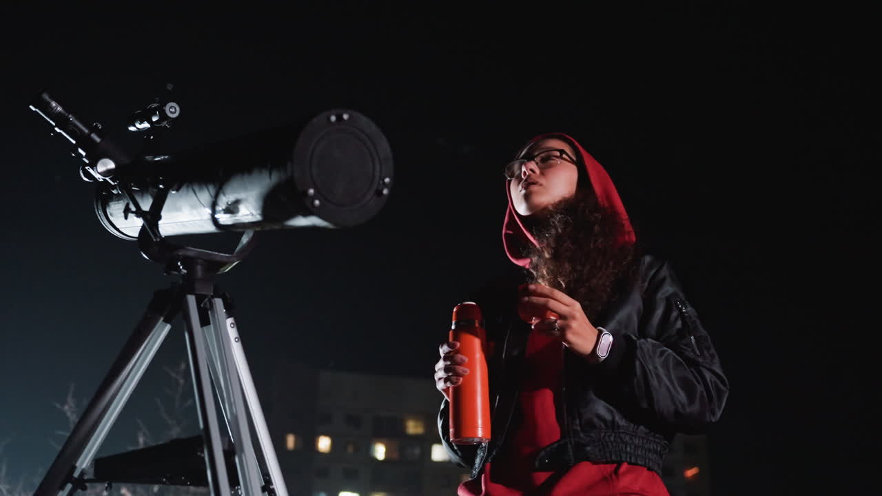 Rooftop astronomer examines telescope at night, hooded figure sips from thermos against city lights, tripodmounted scope aimed at stars, cinematic lowkey lighting and contemplative atmosphere
