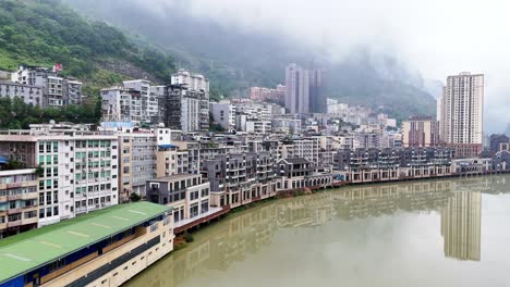 mountain-village-skyline-shot-of-Yunnan,-Yanjin-village-from-river,-apartment-buildings-and-chinese-homes,-water-reflection
