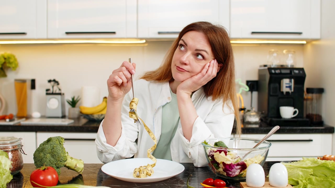 Donna di mezza età sconvolta con un metro a nastro in cucina che riflette su dieta dimagrante, forza di volontà e forma fisica