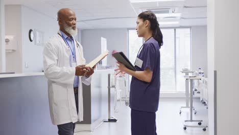 Diverse-male-and-female-doctors-holding-clipboard-and-tablet,-talking-at-hospital