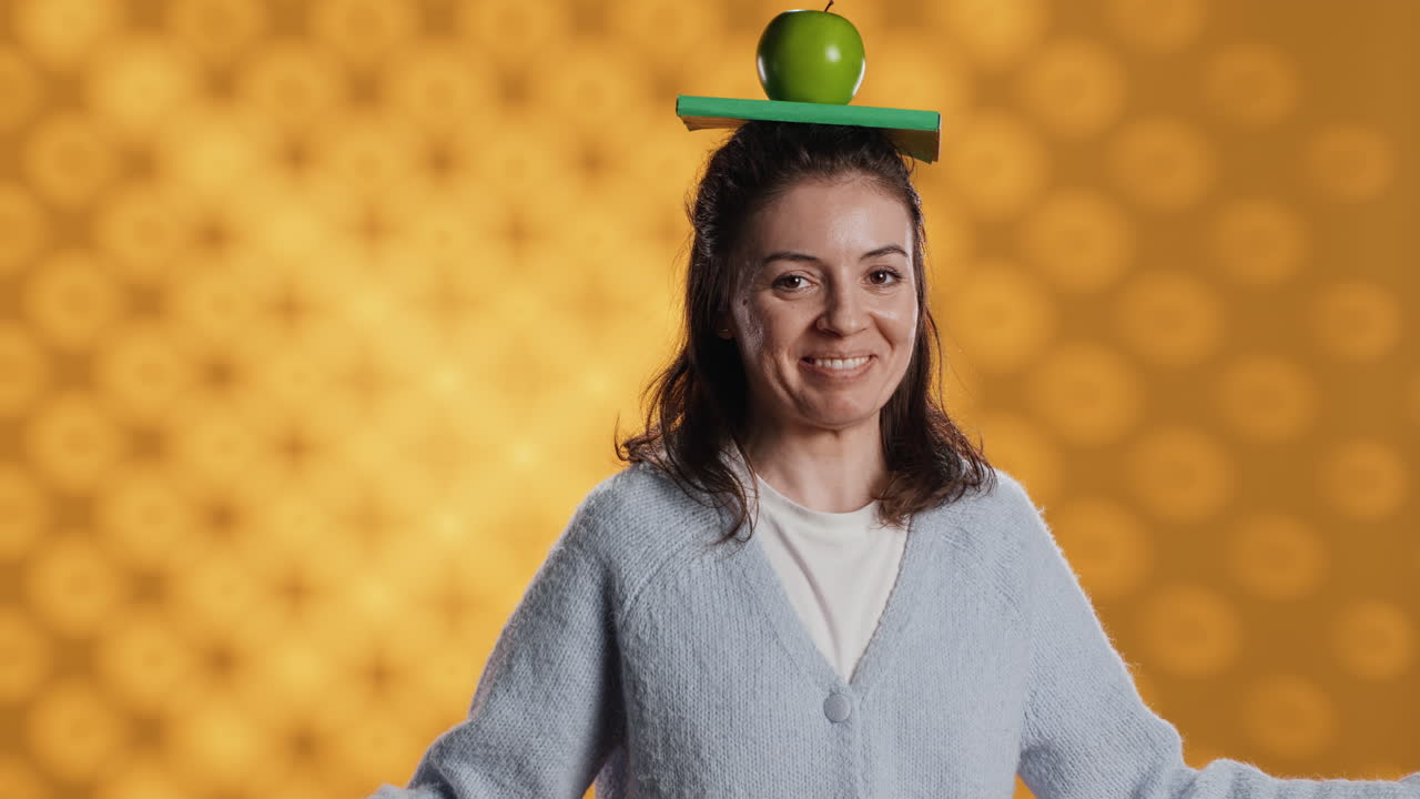 mujer equilibrando libro y manzana en la cabeza, actuando zany, fondo de estudio