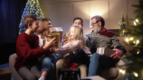 Happy-Family-Gathered-Together-At-Cozy-Home-Near-Decorated-Glowing-Xmas-Tree-Smiling-And-Giving-Xmas-Gifts-To-Each-Other-On-Christmas-Eve-In-Evening