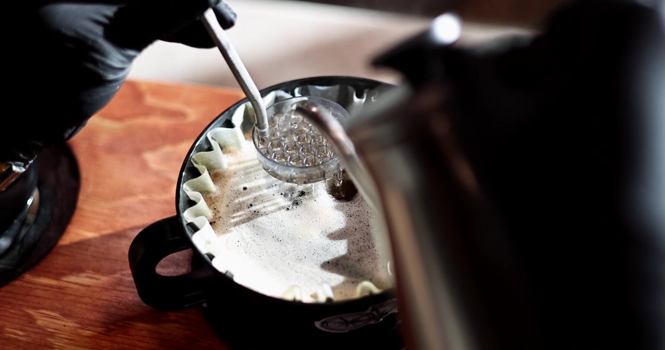 Mano guantata mescola caffè pour over in fioritura mentre l'acqua calda scorre da un bollitore a collo d'oca all'interno, POV