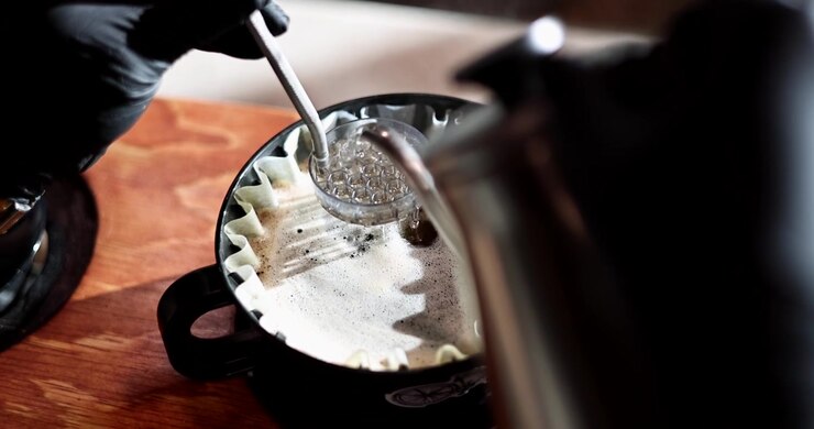 Mano guantata mescola caffè pour over in fioritura mentre l'acqua calda scorre da un bollitore a collo d'oca all'interno, POV