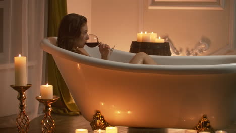 woman bathing with pleasure lying down in the tub with foam and drinking red wine spending time in luxury spa resort