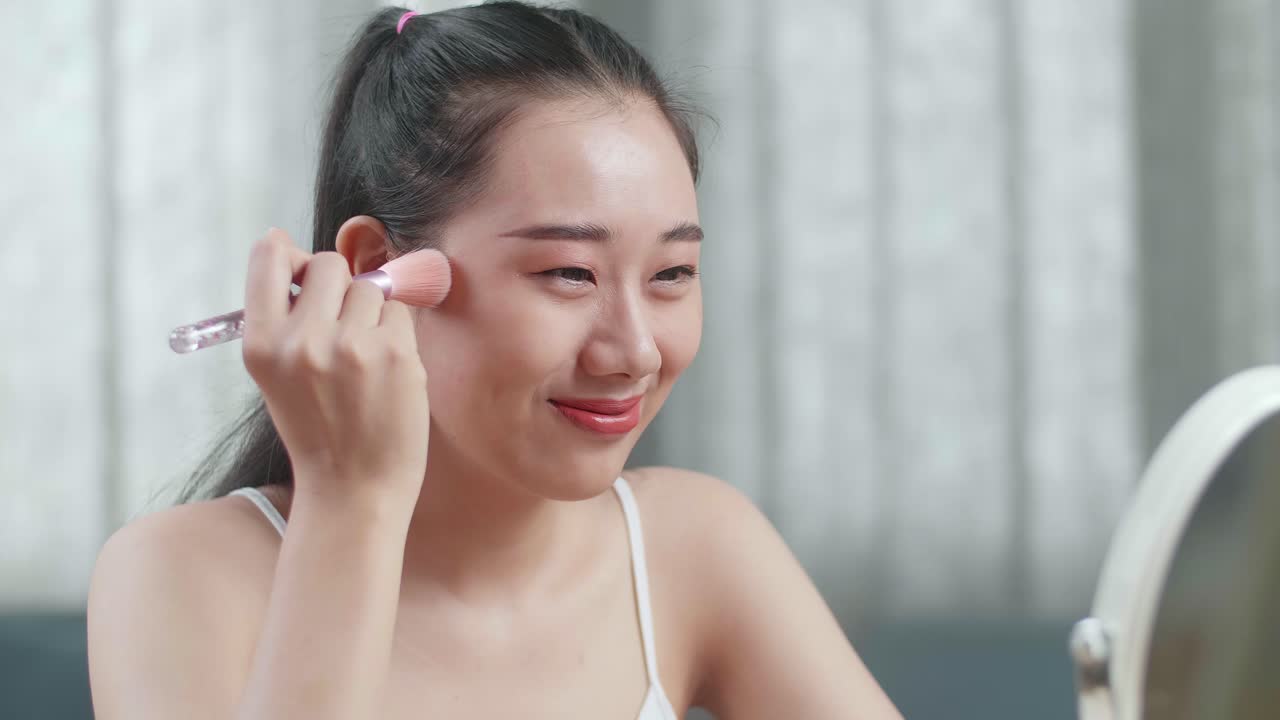 아름다운 젊은 아시아 여성이 거울을 바라보며 을 고 집에서 메이크업하는 동안 미소 짓고 있습니다.
