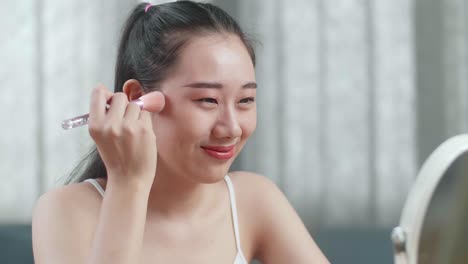 아름다운 젊은 아시아 여성이 거울을 바라보며 을 고 집에서 메이크업하는 동안 미소 짓고 있습니다.