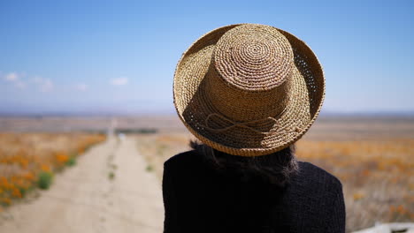 an old woman in summer hat looking down an abandoned dirt road in a field of orange california poppy flowers slow motion