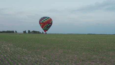 passenger riding colorful hot air balloon drifting low over green farm land at sunset sky with glowing flame illuminating peaceful horizon and soft clouds