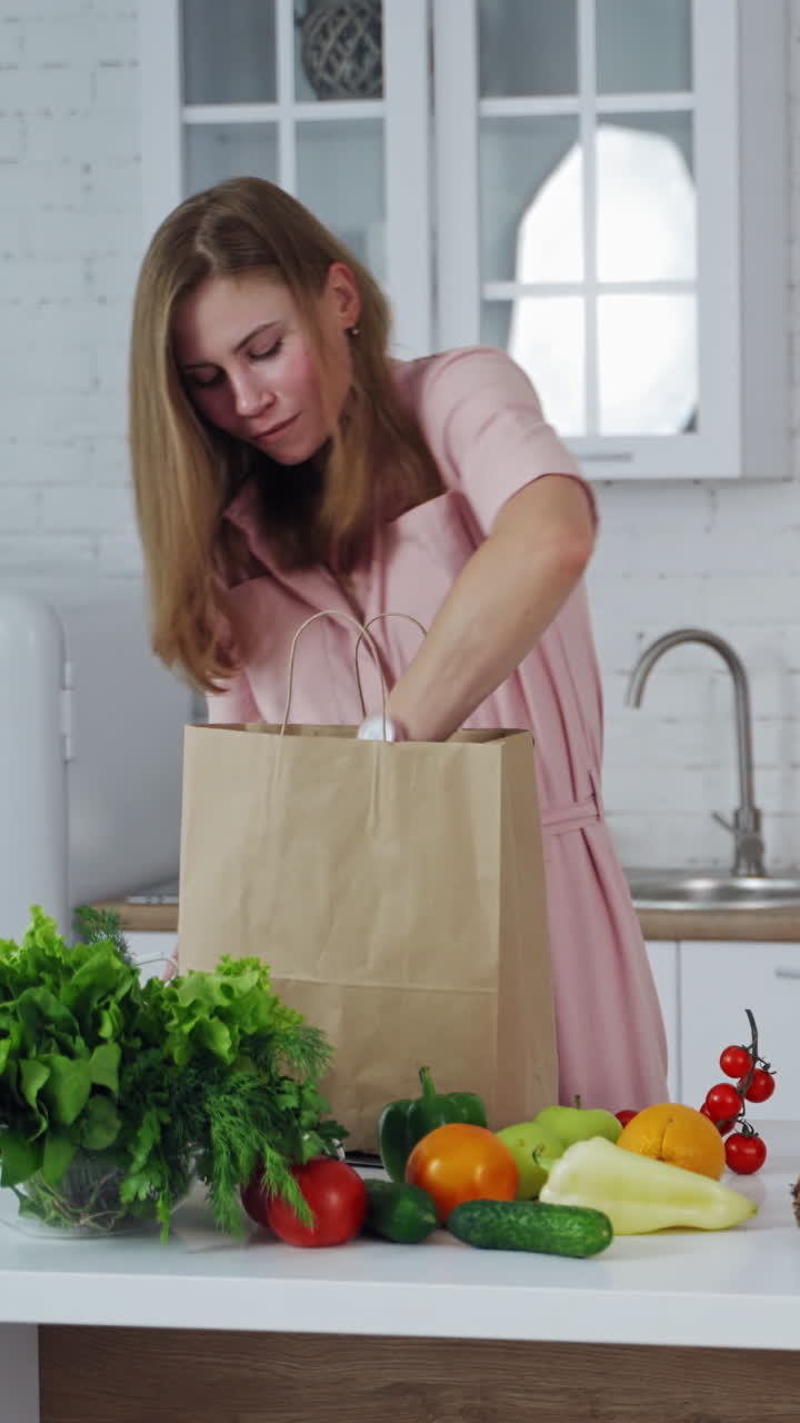 Happy housewife with fresh fruit and vegetables in the kitchen. Young woman unpack organic food from paper bag at home. Vertical video