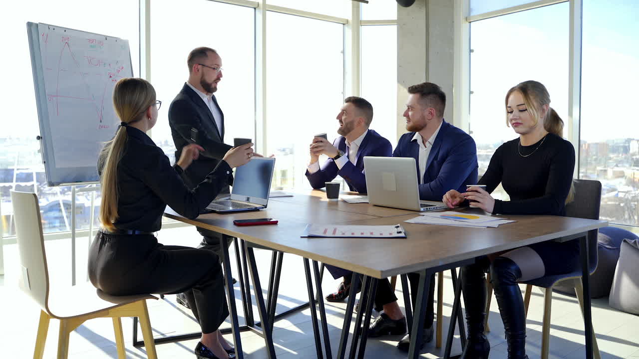Office room with business partners. Group of young entrepreneurs sitting at the table and listen to a cheerful businessman.