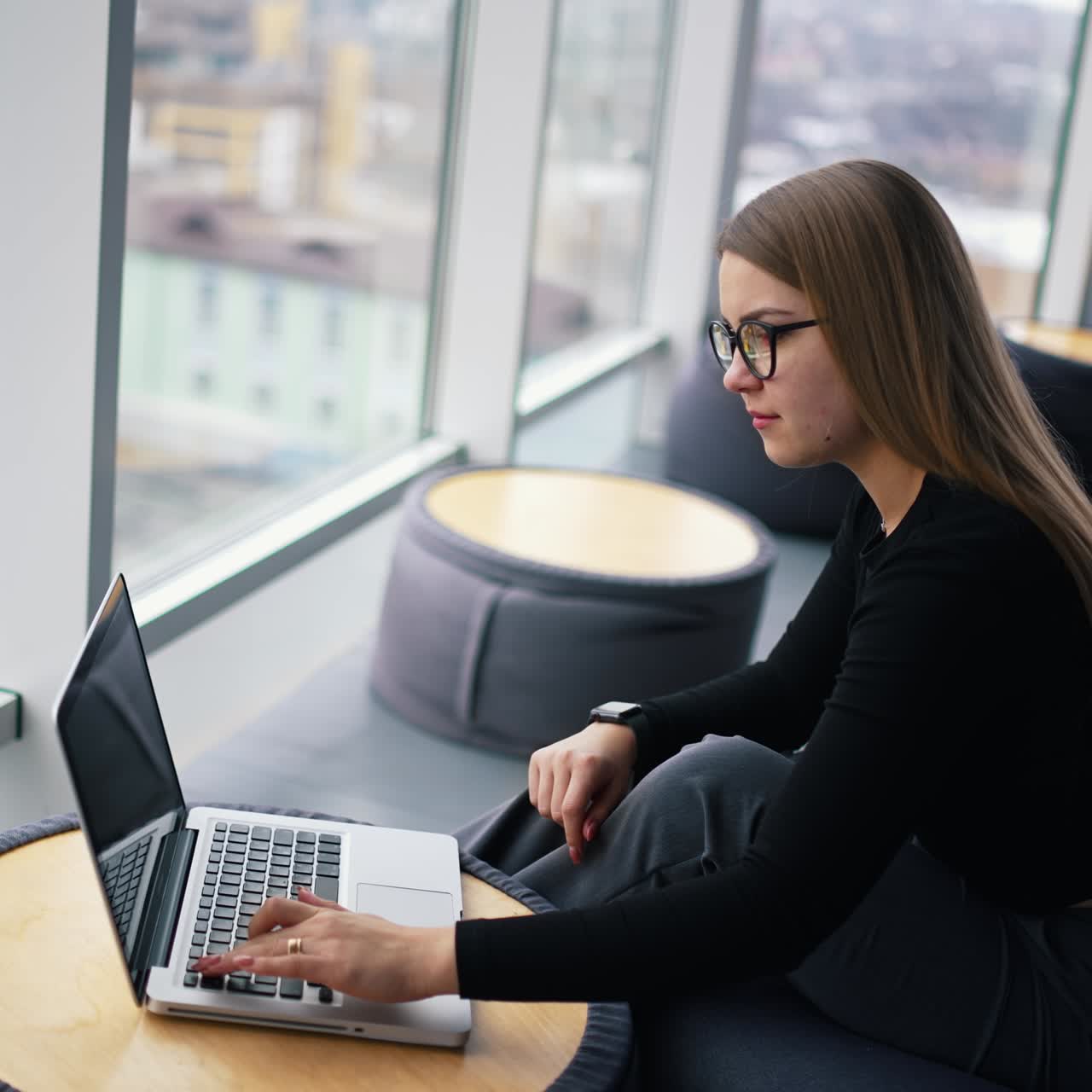Giovane donna elegante in un interno d'ufficio. Bella donna d'affari con gli occhiali che lavora al computer portatile vicino alla finestra con vista sulla città.