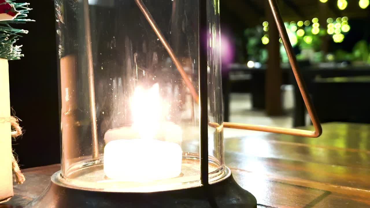 A candle glows warmly inside a glass holder, casting reflections on a wooden table surface.