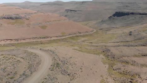 Toma-Aérea-Siguiendo-A-Dos-Ciclistas-De-Montaña-Subiendo-Un-Paso-De-Montaña-De-Camino-De-Ripio