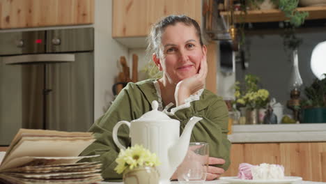 portrait view of woman in green dress sitting at kitchen table smiling warmly into camera with hand on jaw, surrounded by teapot, dessert plate, glass mug, open book and flowers