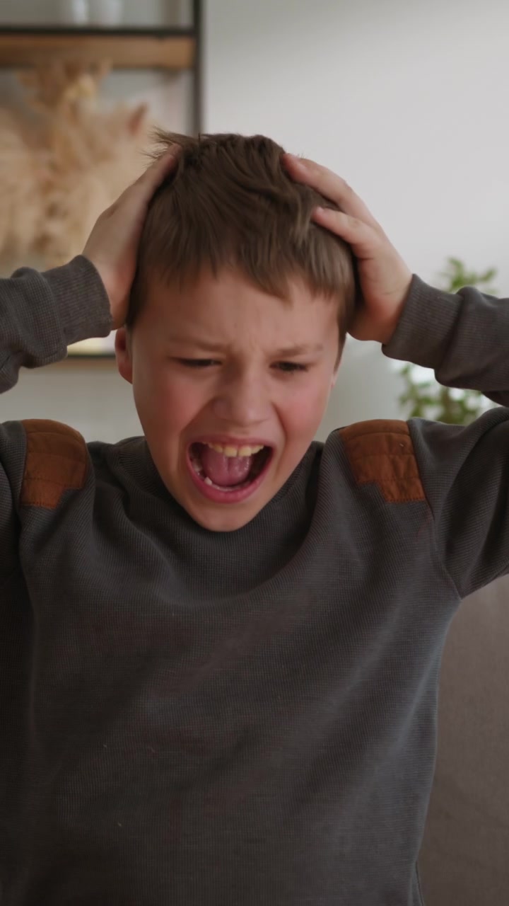 Young boy expresses extreme frustration and distress by screaming with hands on head indoors