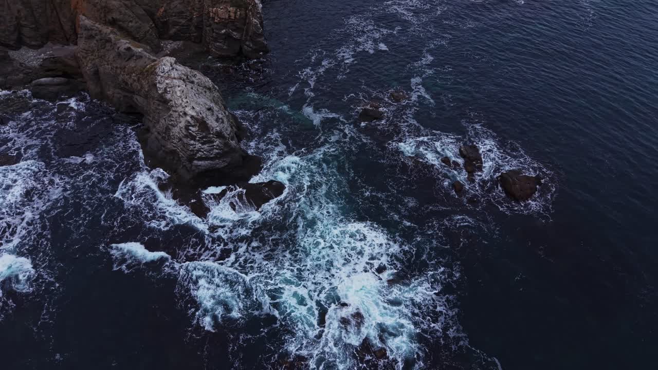 Waves crash against rocks in ocean near rocky coastline at dusk
