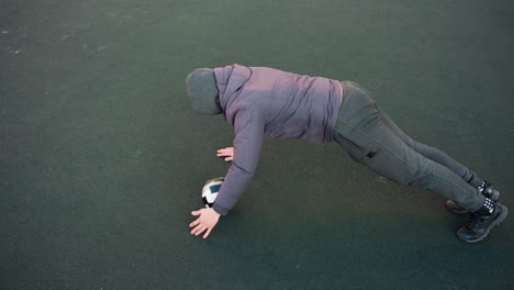 overhead view of athlete performing push-ups with alternating hands on soccer ball demonstrating strength, control, and agility during outdoor training session on plain green surface