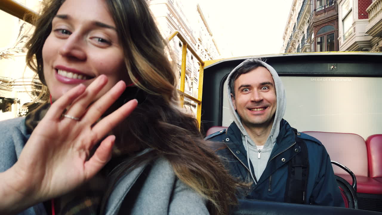 A couple enjoys their day out in the city, riding on the top deck of an open bus. They share smiles and take in the sights while listening to music, feeling the warm sun