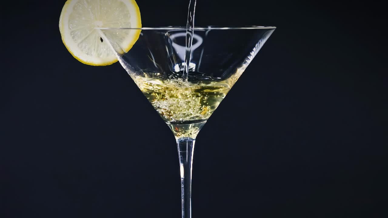 Una fetta di limone decora un bicchiere da martini mentre viene versato del liquido, su uno sfondo scuro.