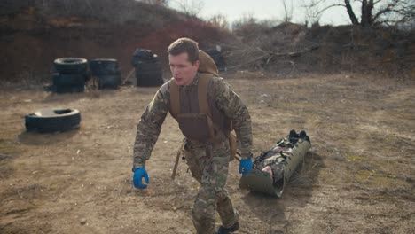 A-tired-confident-young-military-man-in-a-camouflage-uniform-and-blue-medical-gloves-drags-an-unconscious-soldier-in-army-camouflage-with-the-help-of-a-special-stretcher-in-the-steppe