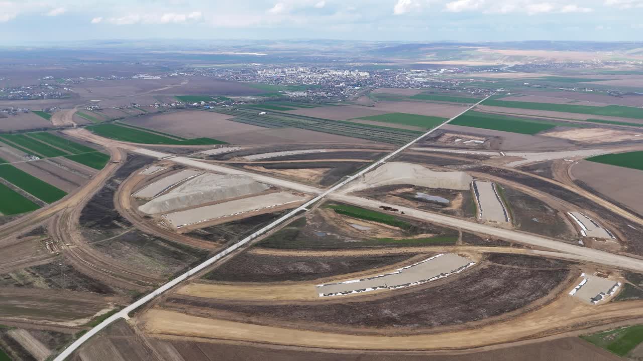 Korkealta otettu dronekuva suuresta moottoritieliittymästä rakenteilla lähellä Pașcania, Romaniaa, kaupungin näkyessä etäällä. Laajempi perspektiivi korostaa mittakaavaa ja tien geometriaa.