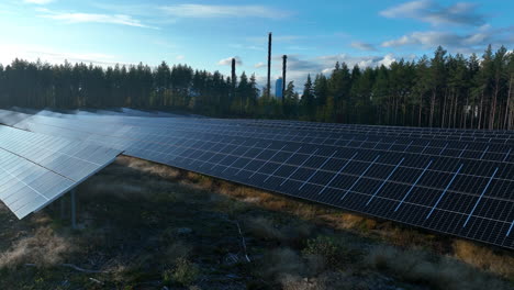 Drone-Volando-Sobre-Un-Campo-Colector-De-Luz-Solar-Con-Fondo-De-Bosque-Y-Chimenea