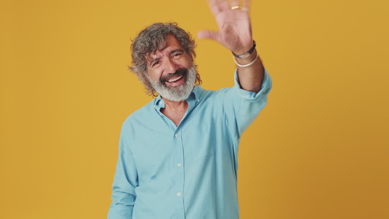 Happy Mature Man with Beard Waving Hello on Yellow Background