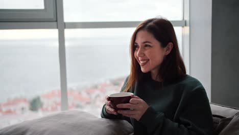 Ein-Glückliches-Brünettes-Mädchen-Freut-Sich,-Während-Es-Auf-Dem-Sofa-Sitzt-Und-Tee-Aus-Einer-Braunen-Tasse-In-Einem-Modernen-Apartment-Mit-Blick-Auf-Das-Meer-Trinkt