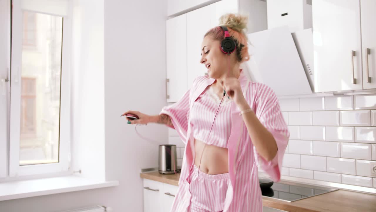 joven feliz con cabello rosa bailando en la cocina con pijama rosa y escuchando música con auriculares